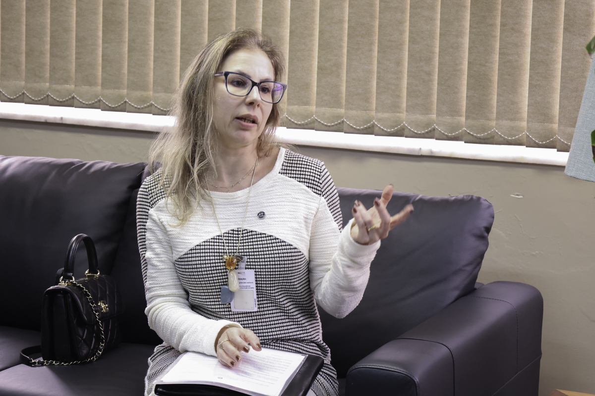 Bianca é branca, loira, de cabelos lisos. Usa óculos de aro grande. Veste blusa branca e preta. Está senta no sofá, segura alguns papeis no colo, e fala olhando para a direita da imagem e gesticulando.