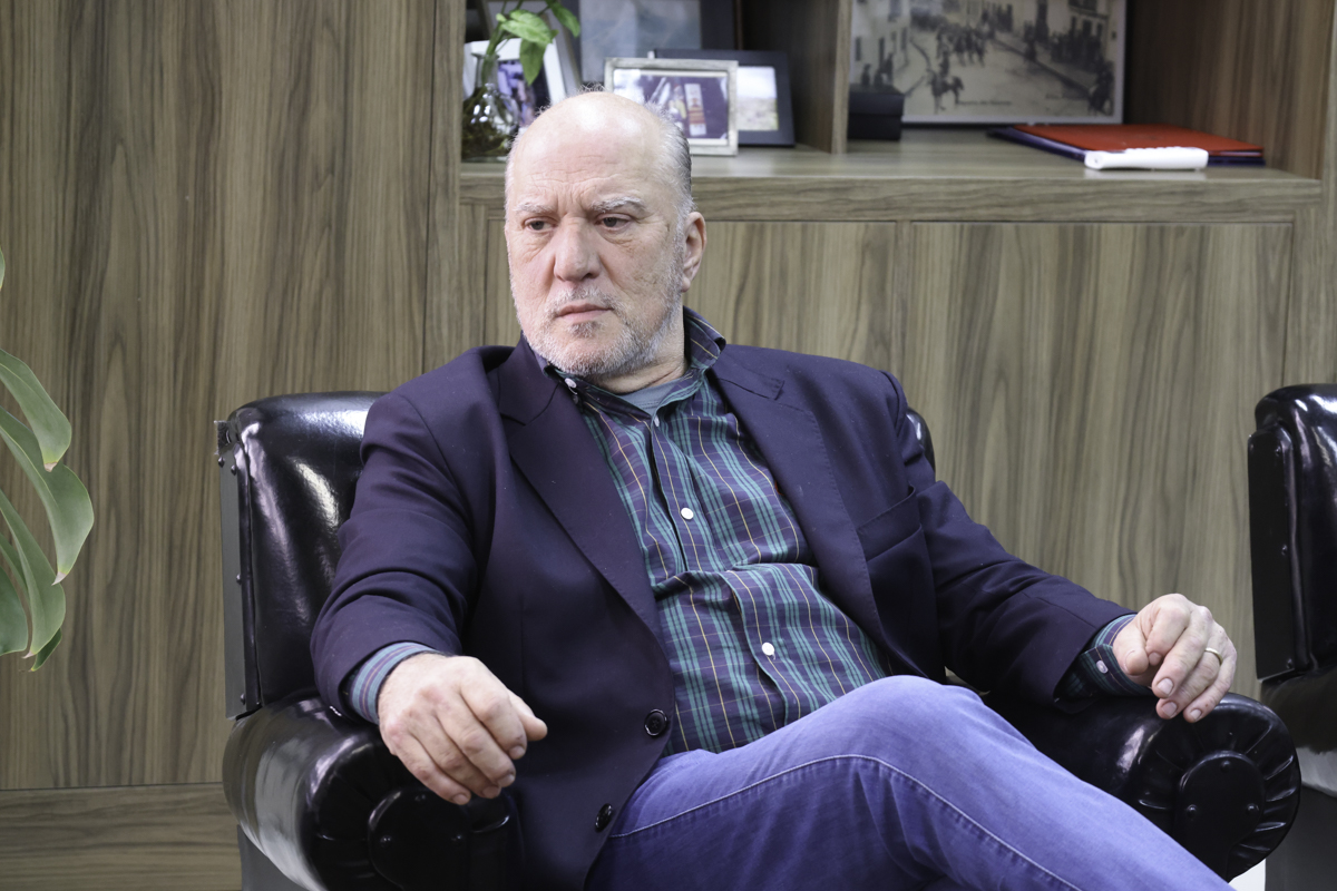 Martins Costa é branco, calvo e grisalho. Usa calça jeans azul, blazer azul escuro e camisa social xadrez em verde e azul. Está sentado em uma poltrona e olha para a esquerda da imagem.