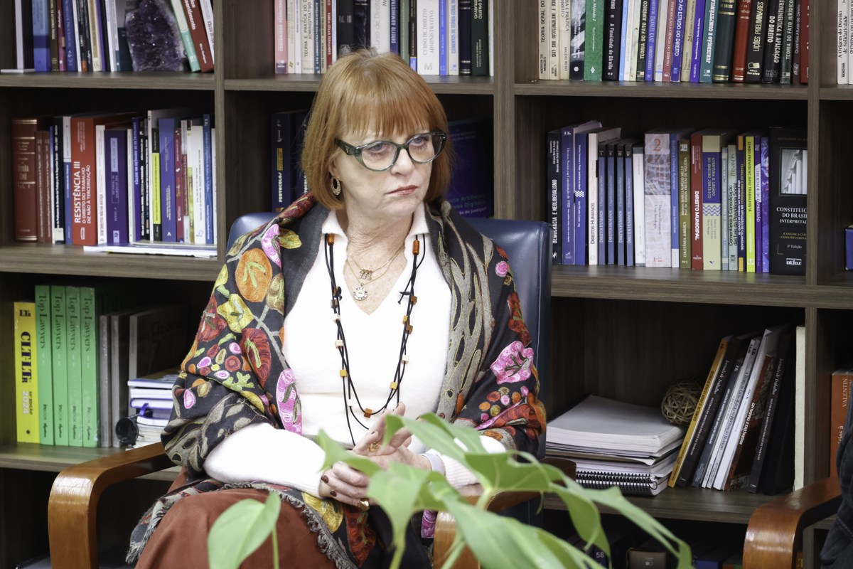 Telesca é branca, ruiva, de cabelos lisos e curto. Usa óculos de aro verde, blusa branca e um xale com estampa florada. Esta olhando para a direita da imagem, atrás dela há uma estante de livros.