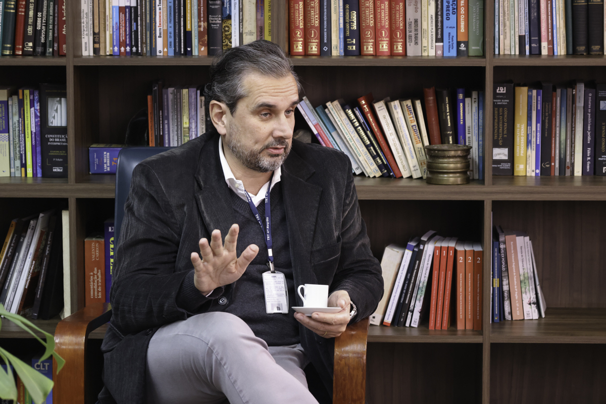 Adolfo é branco, grisalho e usa barba curta. Veste calça cinza, camisa branca, blusão cinza e blazer escuro. Fala e gesticula olhando para a direita da imagem.  Atrás dele há uma estante de livros.