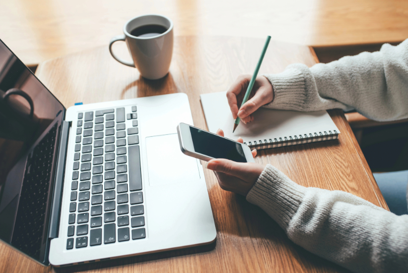 Mãos femininas anotam em um caderno e seguram celular sobre mesa de trabalho na qual há um notebook e uma xícara de café.