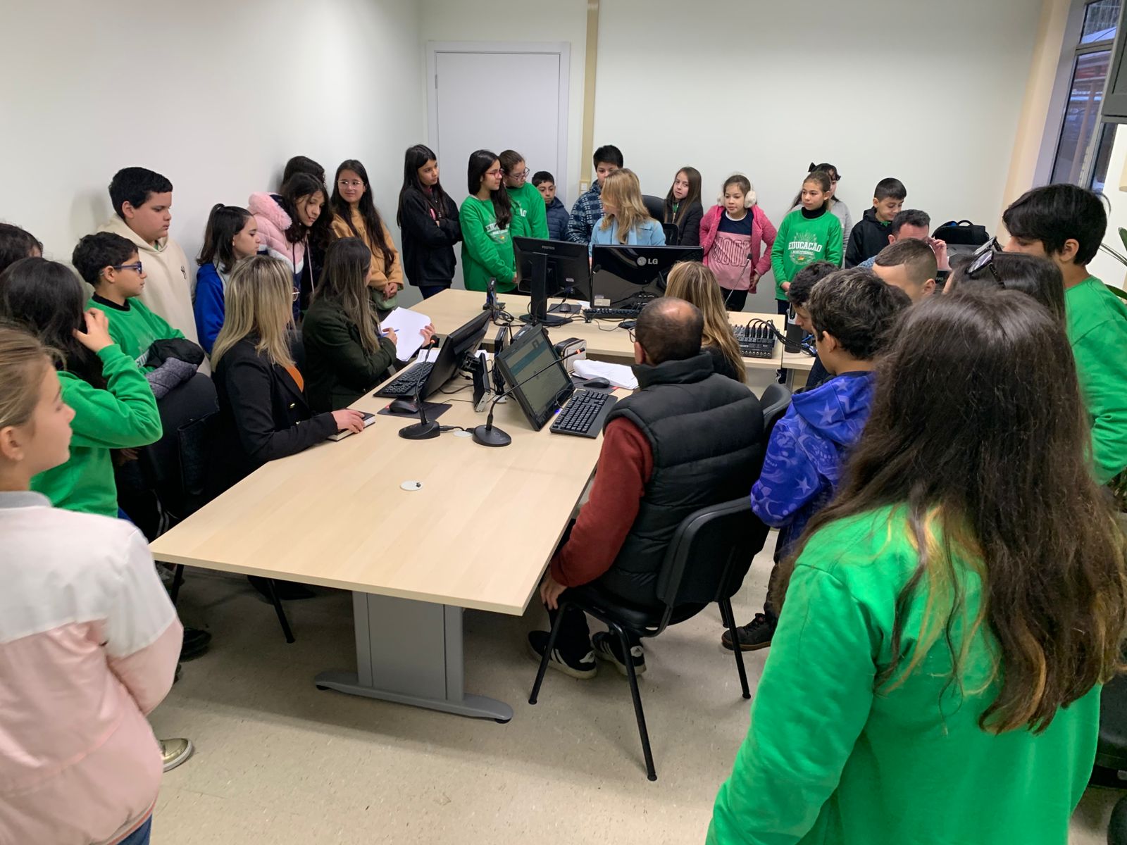 Vários alunos do 5 ano estão em uma sala com computadores, observando servidores em atividade. Estão atentos, ao redor da mesa, acompanhando explicações sobre o funcionamento interno da Justiça do Trabalho.