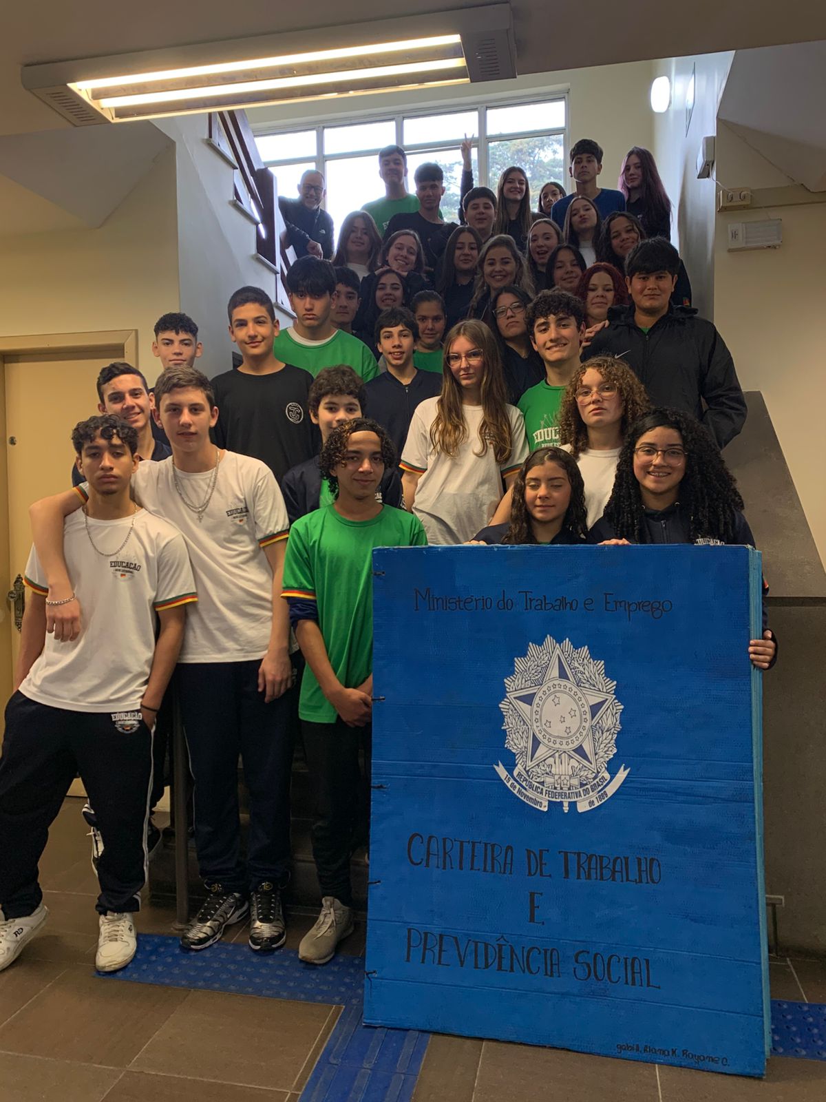 Um grupo de estudantes posa em escadaria interna ao lado de uma réplica gigante da Carteira de Trabalho e Previdência Social.