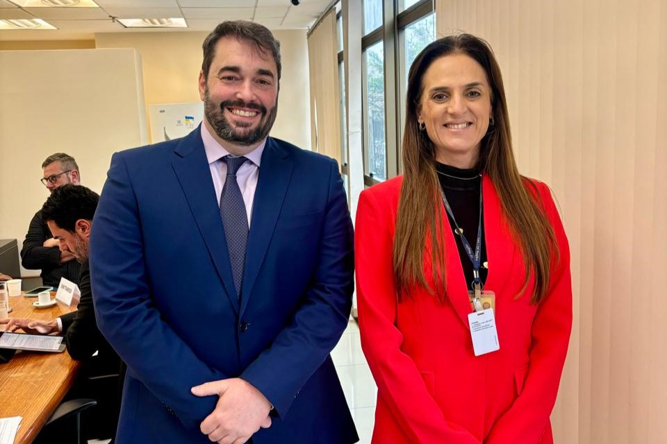 Alexandre e Tamara posam para a foto de pé sorrindo. Ele é branco, de cabelo e barba escura, veste terno azul marinho, camisa rosa e gravata cinza. Ela é branca, de cabelos castanhos compridos. Usa blusa preta, casaco vermelho e um crachá de visitante. Ao fundo aparecem duas pessoas sentadas em uma mesa de negociação.