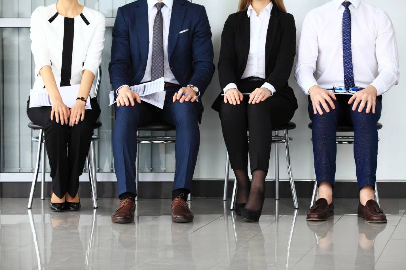 Quatro pessoas sentadas uma ao lado da outra, em provável espera de entrevista de emprego. São dois homens e duas mulheres e não há imagem dos rostos.