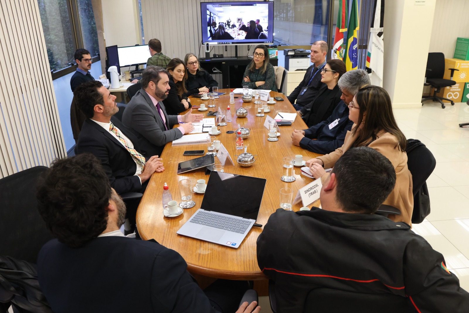 Participantes na mesa de negociação. O vice-presidente está falando, no centro do lado esquerdo. Ao fundo aparece um telão e bandeiras do RS, Brasil e Justiça do Trabalho.