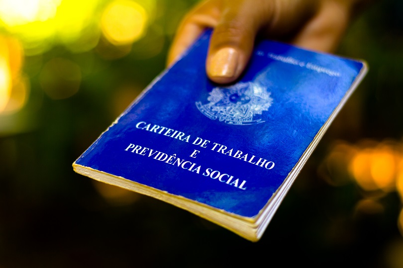 Foto ilustrativa mostra mão de uma pessoa segurando uma Carteira de Trabalho. A pessoa tem a pele morena e a CTPS tem a cor azul.