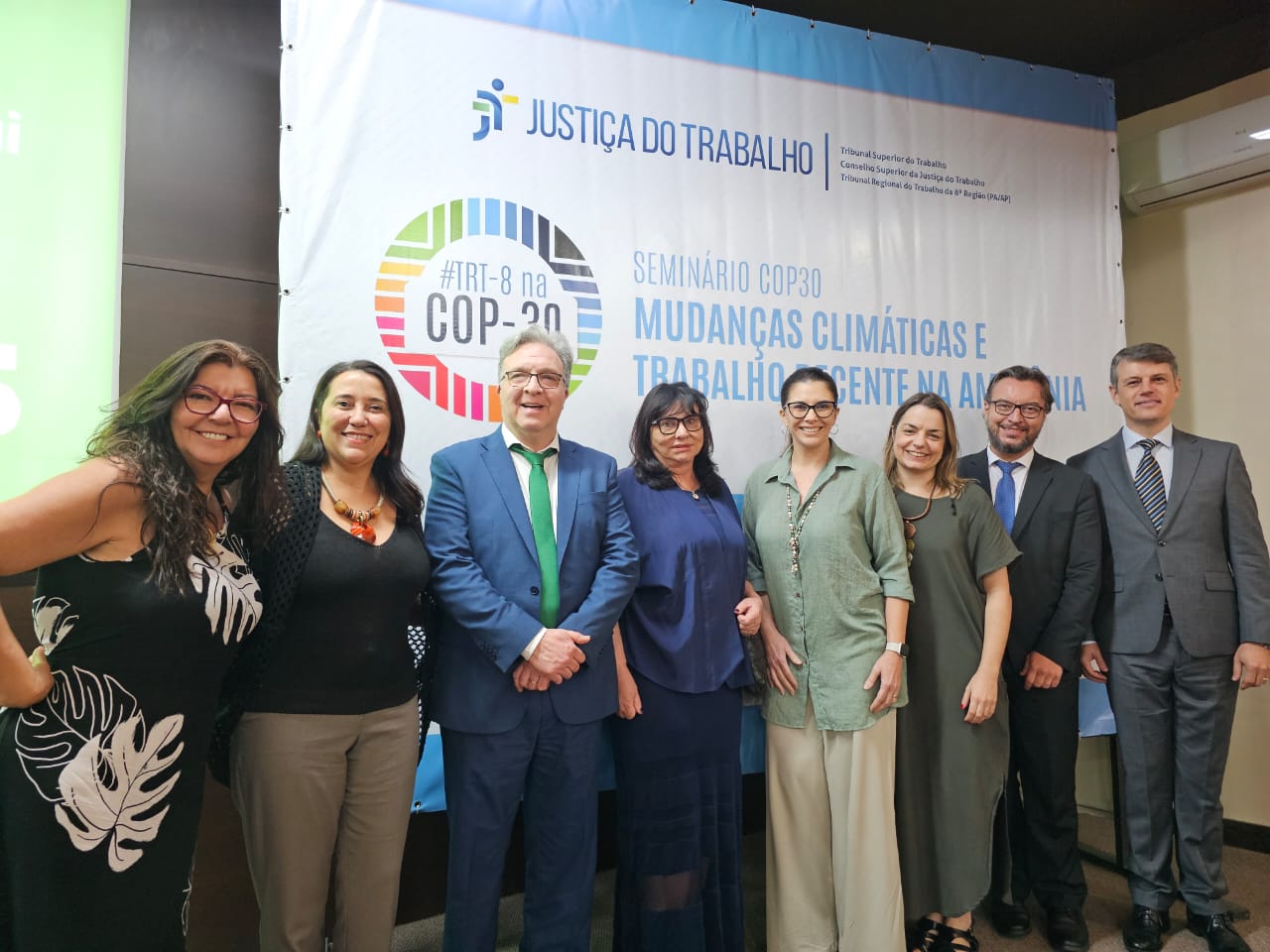 Grupo de oito pessoas posa sorridente diante de painel do Seminário “Mudanças Climáticas e Trabalho Decente na Amazônia”.