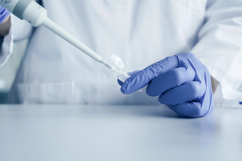 Pessoa com jaleco branco e luvas azuis manuseia pipeta em laboratório, transferindo líquido para pequeno tubo, em possível procedimento de fertilização in vitro.
