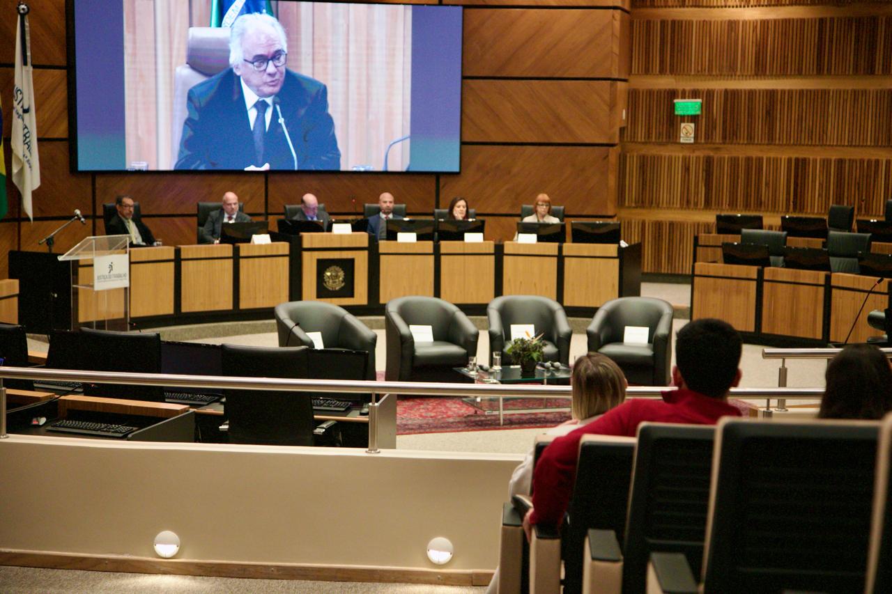 A foto mostra o ministro godinho discursando e sua imagem aparecendo num telão.