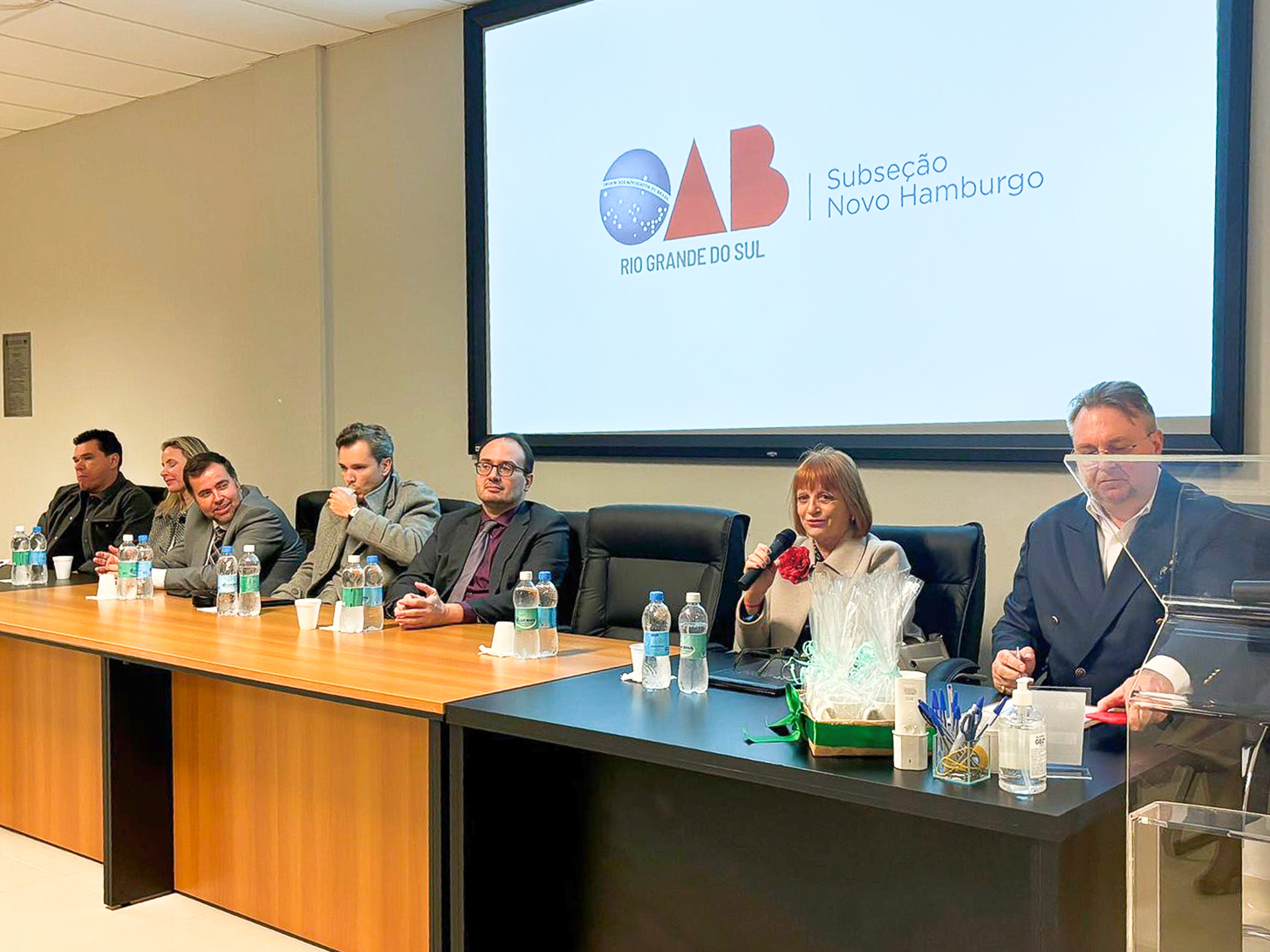 Mesa de abertura de evento na OAB/RS - Subseção Novo Hamburgo, com sete magistrados sentados lado a lado. Ao centro, a desembargadora Telesca segura um microfone enquanto fala. À direita, há um telão projetando o logotipo da OAB do Rio Grande do Sul.