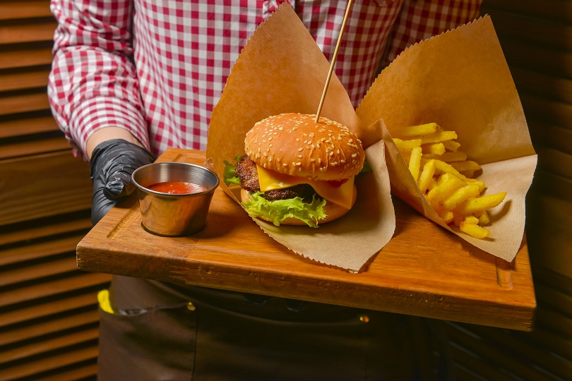Homem segura bandeja com hambúrguer, batatas fritas e ketchup. Ele usa camisa xadrez vermelha e branca e calça marrom. Não há imagem do rosto.