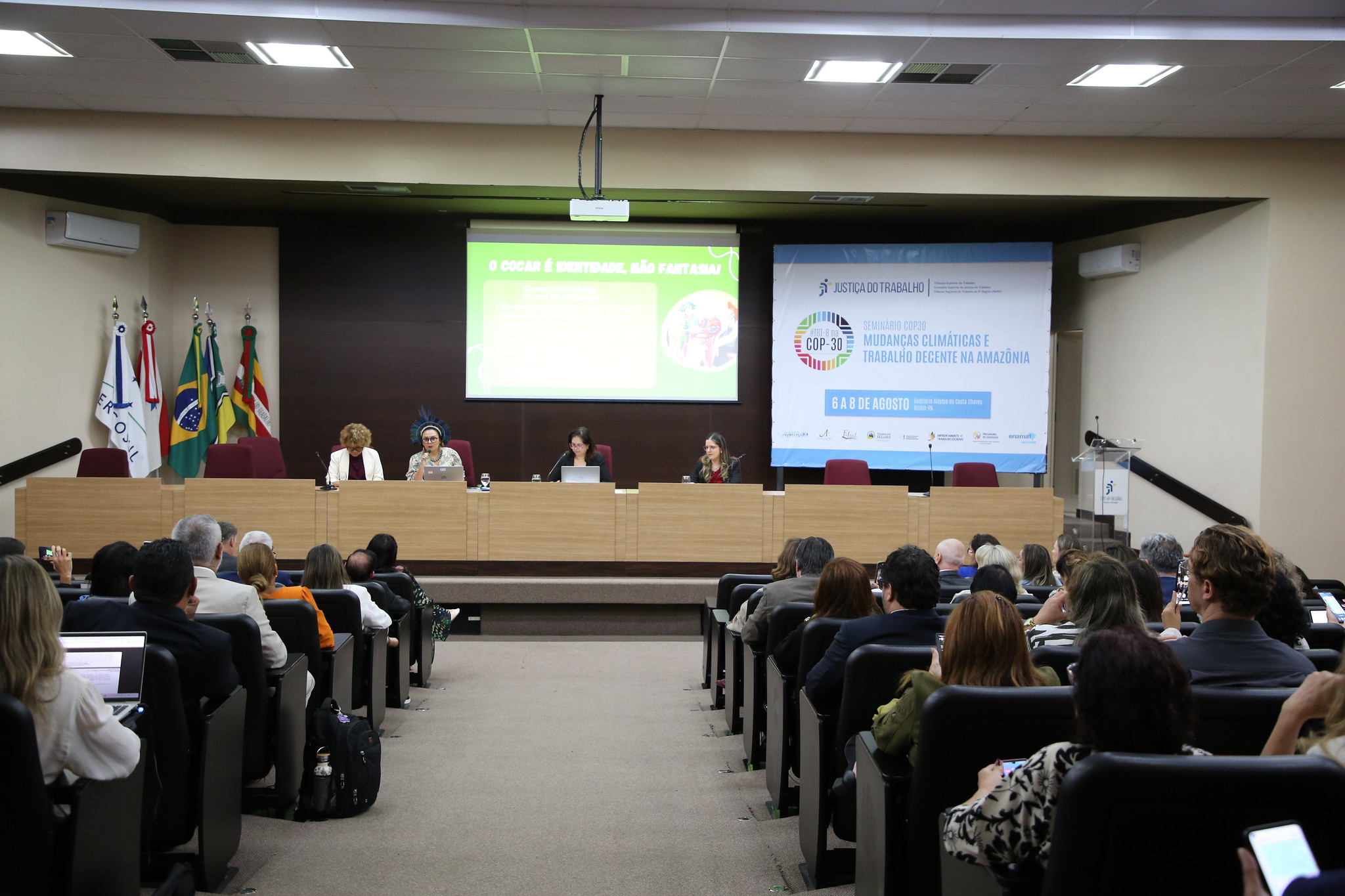 Foto do evento, em ângulo aberto, mostrando público de costas e mesa ao fundo