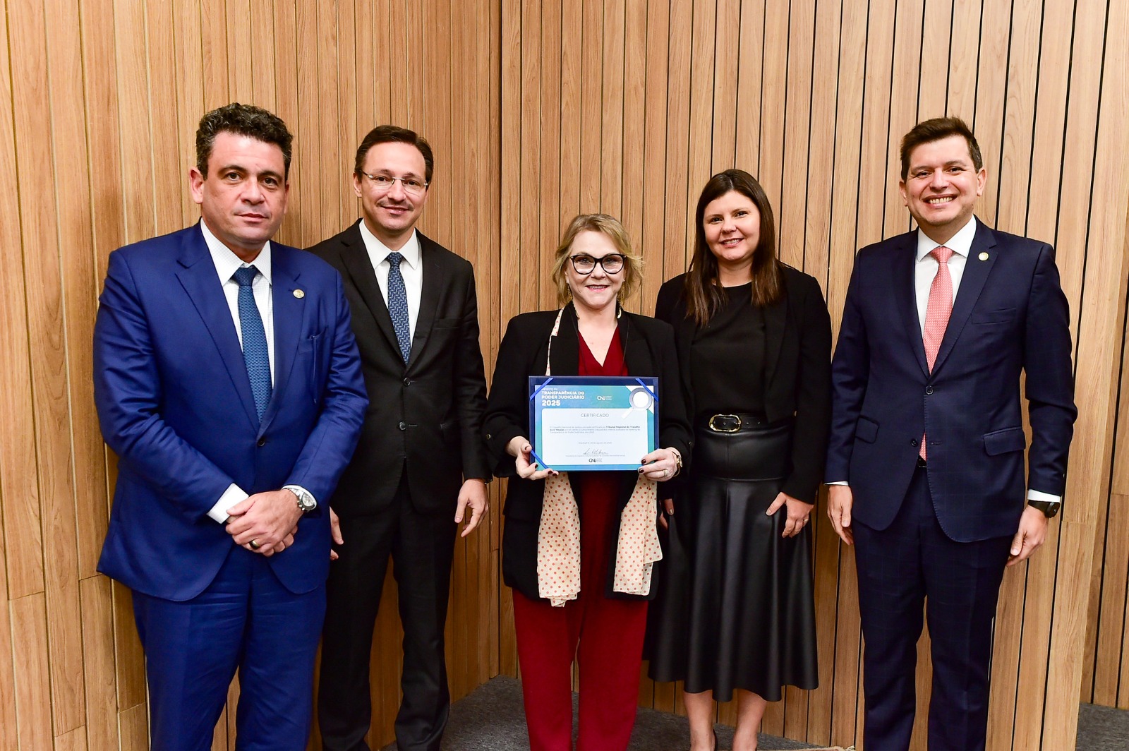 Cinco pessoas posam sorrindo para a foto. Laís est no centro segurando o certificado. É branca, loira, e usa vestido vermelho com casaco preto. 