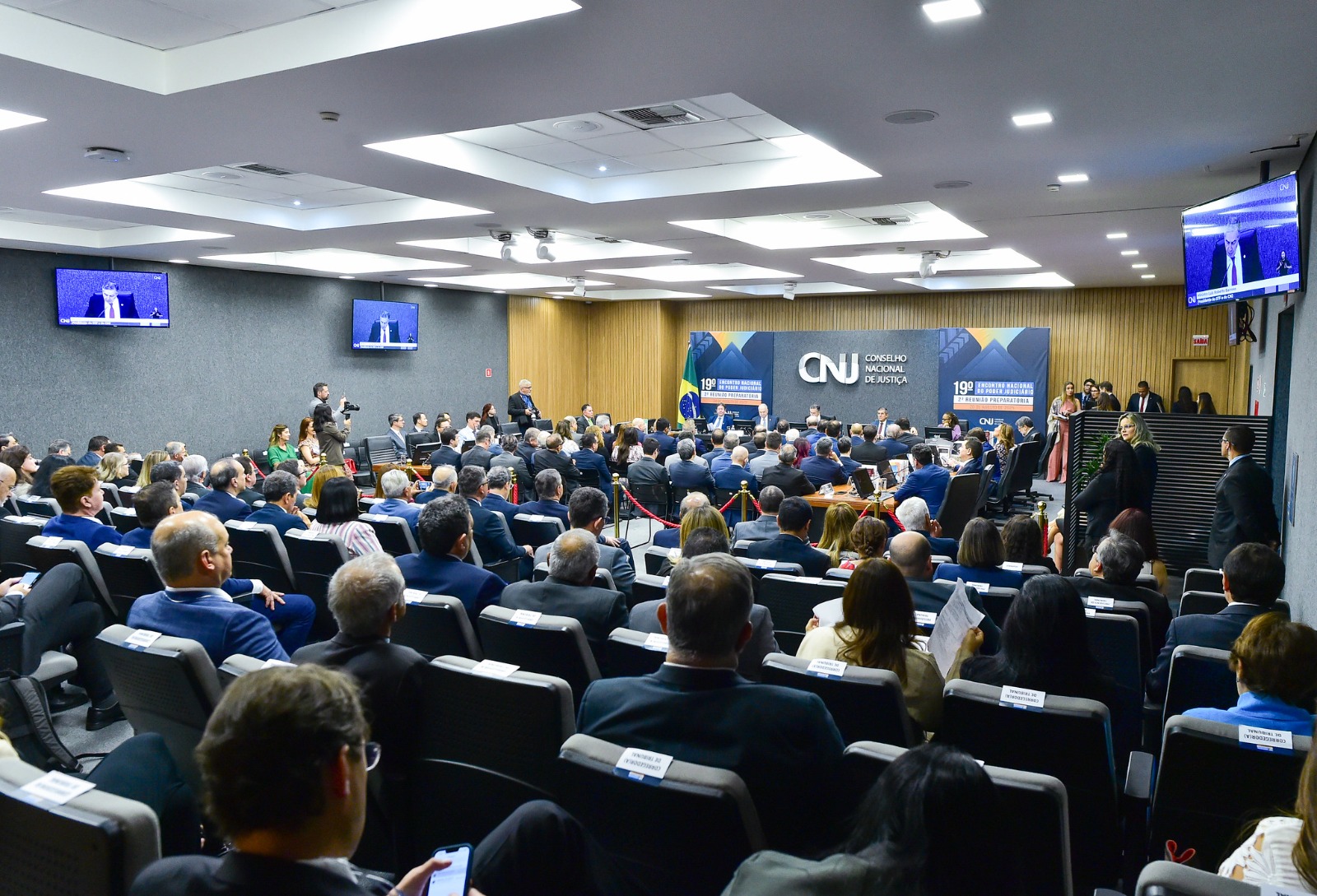 Foto de uma sala ampla. Ao fundo aparece a mesa oficial e o logo do CNJ. O público está de costas em primeiro plano. 