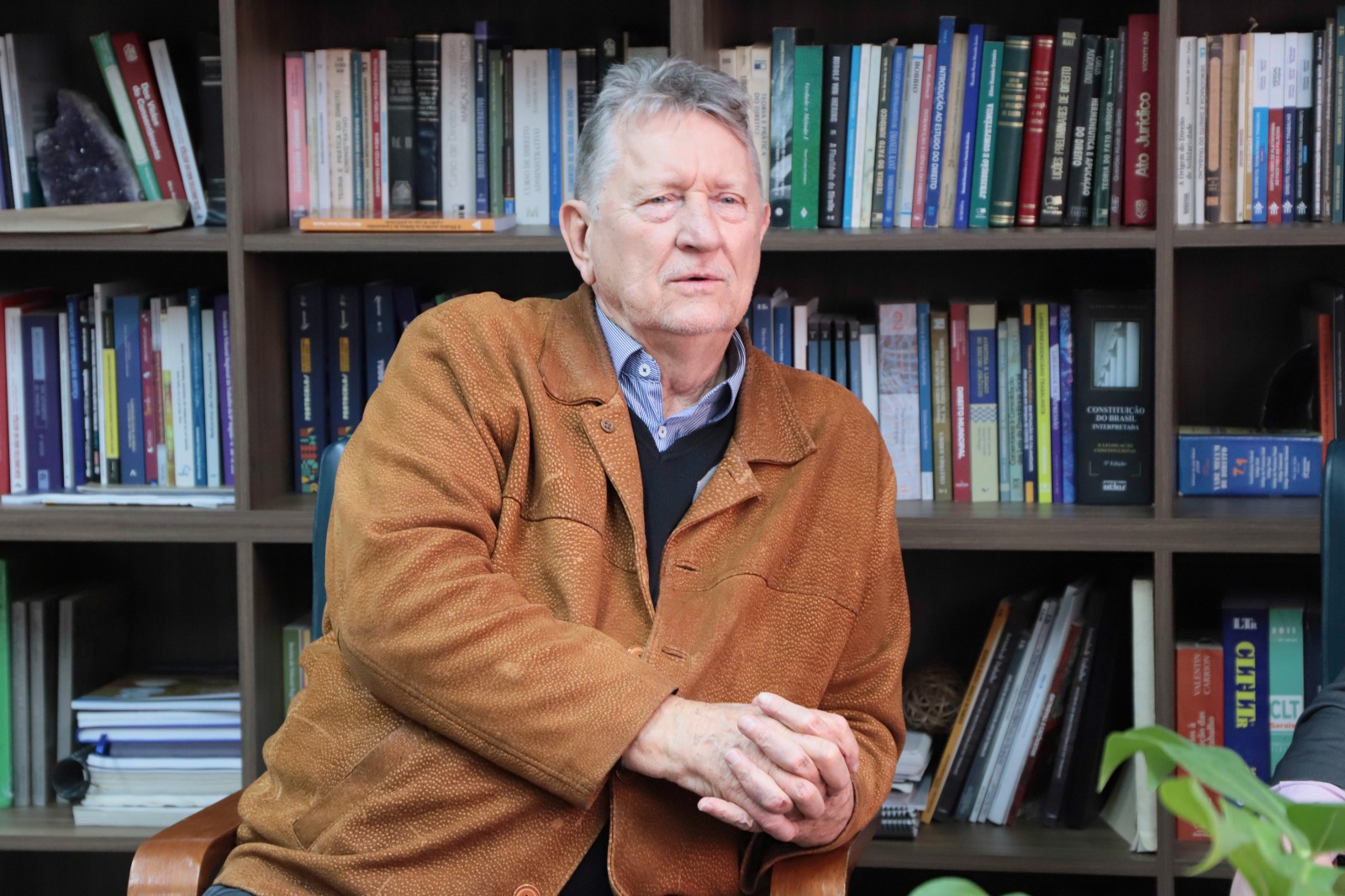 Leonid é branco e grisalho. Usa um casaco de couro, blusão preto e camisa social azul. Ele está sentado, falando e com as mãos entrelaçadas. Ao fundo aparece a estante com livros.