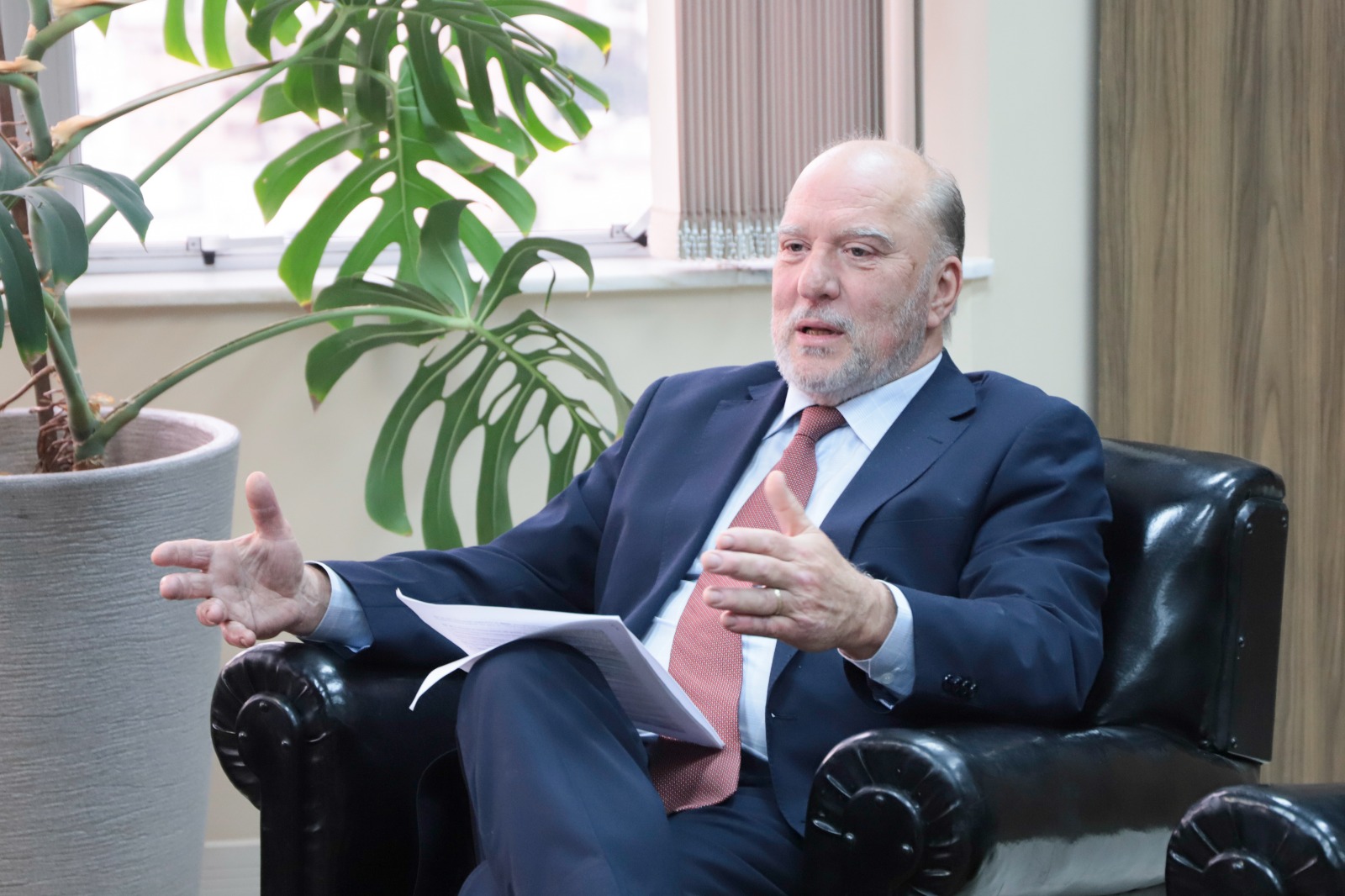 Martins Costa é branco, calvo e grisalho. Ele está sentado em uma poltrona, falando e gesticulando. Usa terno azul marinho, camisa branca e gravata rosa. A seu lado aparece uma planta em um vaso e ao fundo a janela aberta.