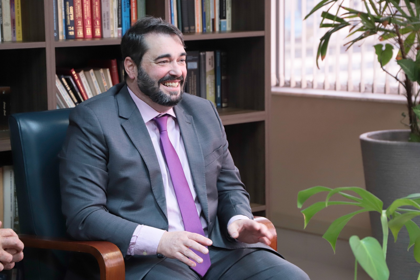 Alexandre é branco e usa cabelo curto escuro e barba escura. Veste terno chumbo com camisa e gravata  em tons de rosa. Ele fala, gesticula e está sorrindo. ao fundo aparece a estante com livros.
