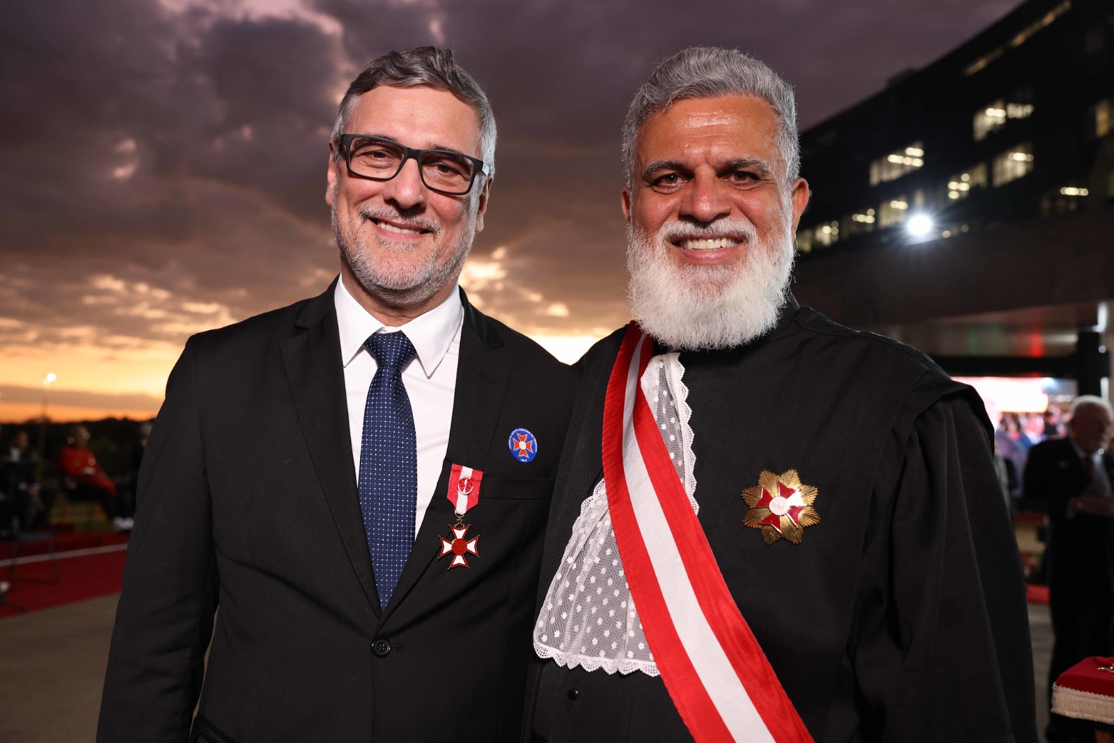 Peixoto é branco, de cabelo e barba grisalha. Veste terno preto, camisa branca, gravata azul e óculos de armação preta. Há duas medalhas na lapela do seu blazer. Lelio é pardo, de cabelo e barba grisalhos. Usa toga, com um faixa vermelha e branca atravessada no peito e uma medalha na lapela. Ao fundo, aparece o prédio do TST e o céu nublado com o pôr do sol no horizonte.