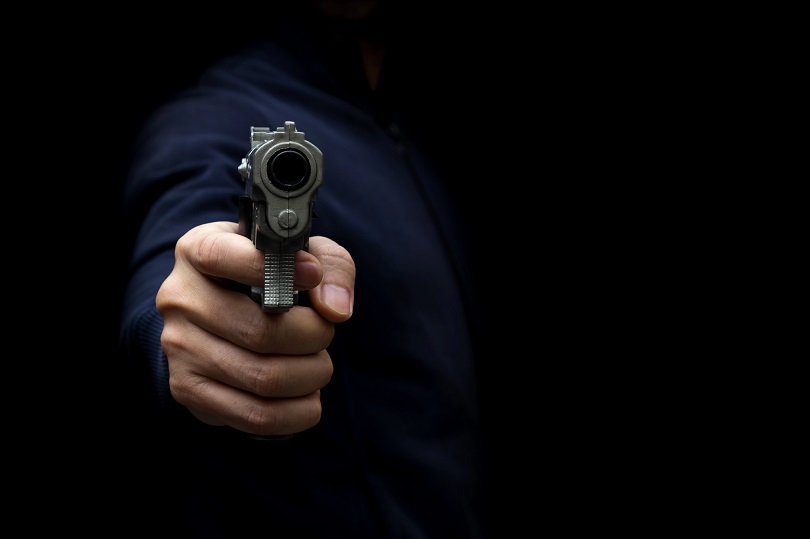 A imagem mostra uma pessoa apontando uma arma de fogo diretamente para a câmera, em um ambiente escuro, transmitindo tensão e ameaça.
