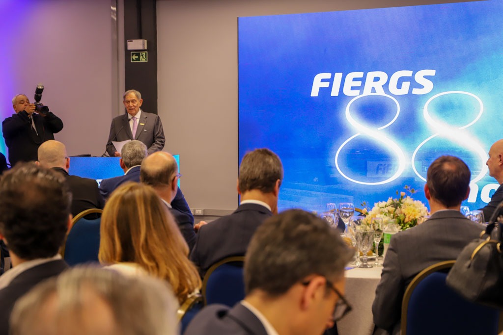 A foto mostra o presidente Claudio Bier de fundo discursando, com público na frente.