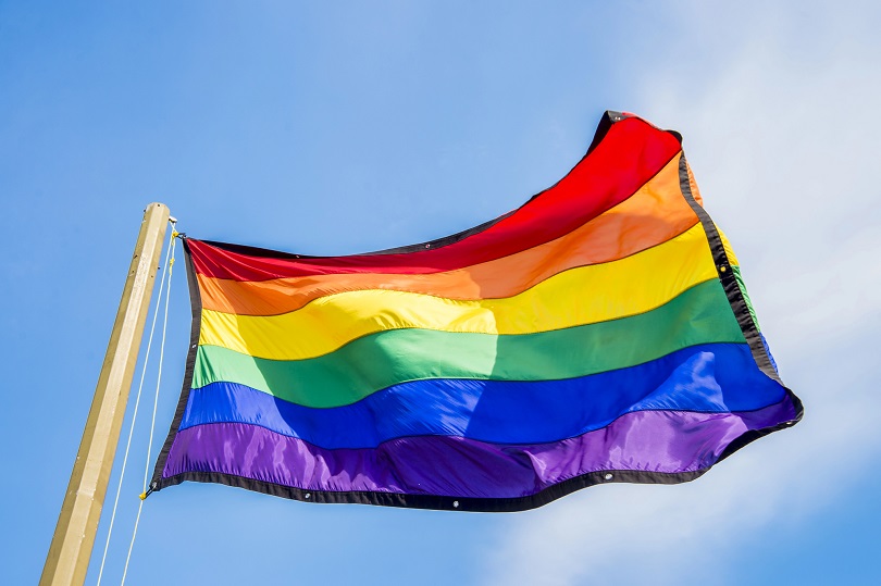 Foto ilustrativa mostra uma bandeira do arco-íris, símbolo do movimento LGBTQIA+, tremulando em um mastro contra o céu azul.