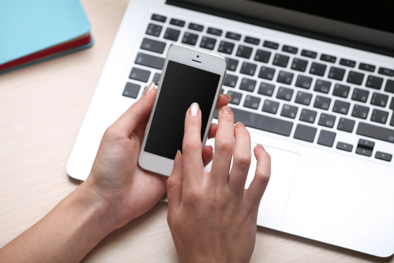Mãos femininas mexem em aparelho celular. Abaixo, há um notebook.