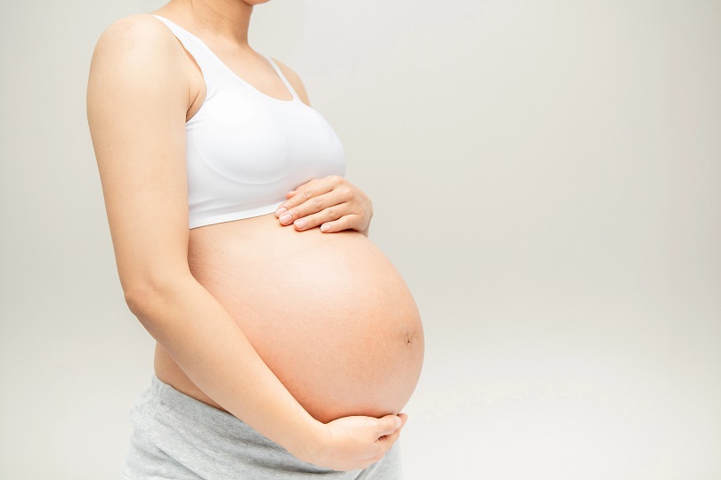 Mulher grávida segura a barriga com as mãos, vestindo top branco e calça cinza, em fundo neutro.
