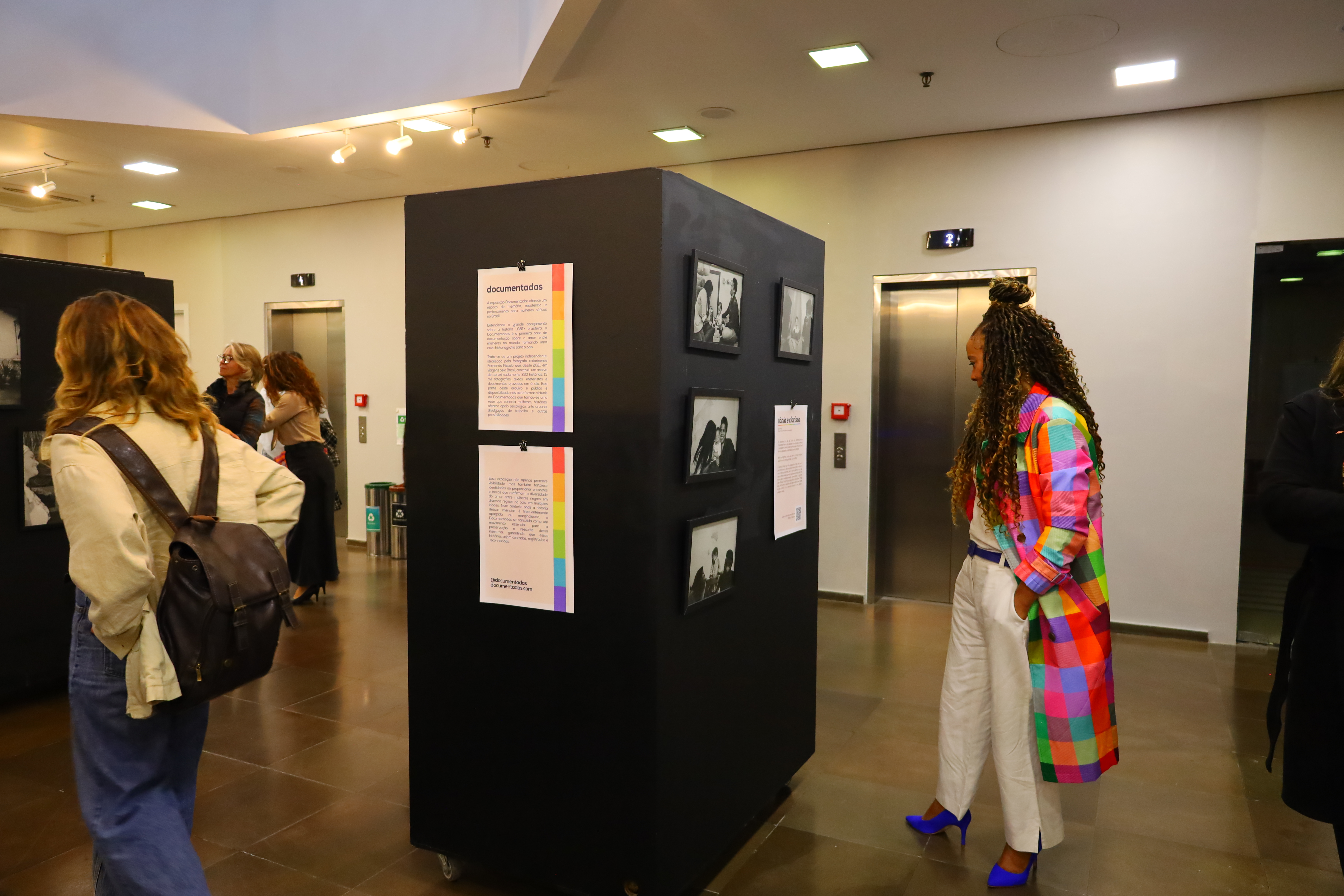 Pessoas circulando, olhando as fotos em painéis pretos. Uma mulher à direita veste casaco colorido quadriculado, calça branca e sapatos azuis, está observando a exposição. 