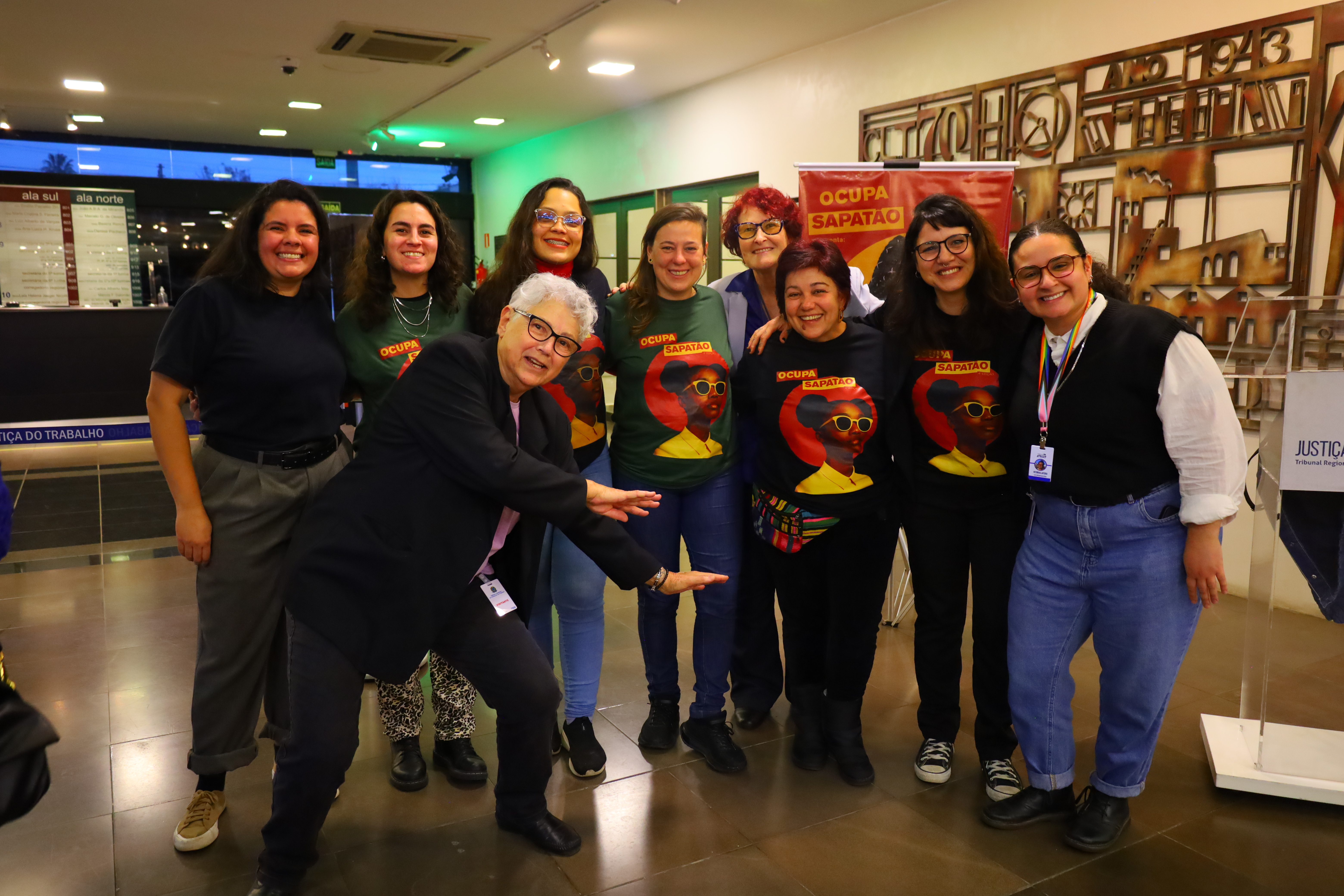 Grupo de pessoas, maioria mulheres, posando para foto. Algumas vestem camisetas pretas com ilustração colorida e esta escrito o nome da ong: “OCUPA SAPATÃO”.