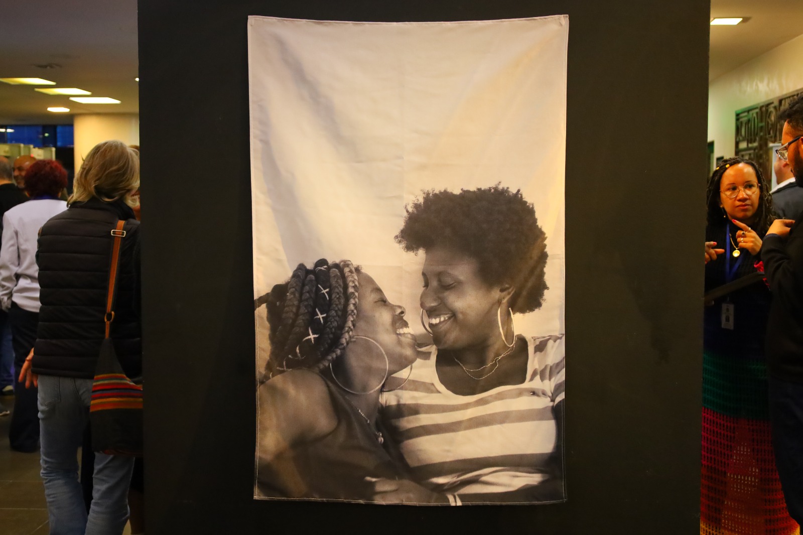 Fotografia em preto e branco de duas mulheres sorrindo e olhando uma para a outra. A imagem está pendurada em um painel preto.