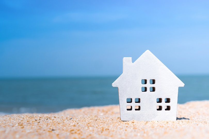 A foto ilustrativa mostra uma pequena maquete branca em formato de casa, colocada sobre a areia da praia. Ao fundo, vê-se o mar e o céu azul.