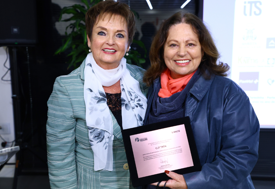 Rejane ao lado de uma mulher sorrindo. A da esquerda veste blazer verde com listras brancas e lenço branco estampado; a da direita (Rejane) usa casaco azul, cachecol laranja e segura a placa de homenagem.