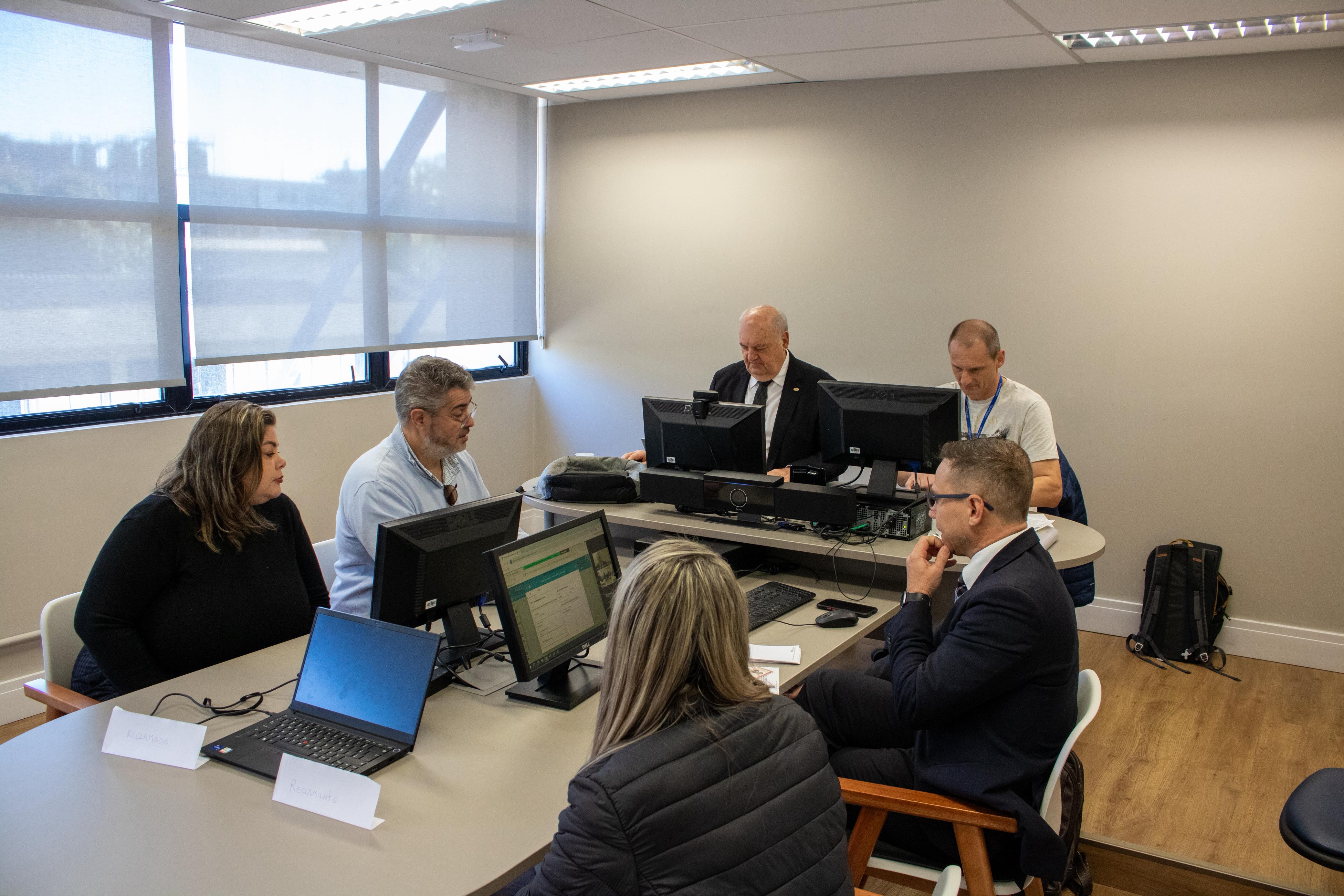 Luiz Colussi conduz audiências trabalhistas com a presença de partes e advogados. Documentos e telas exibem os processos, enquanto os participantes acompanham com atenção.