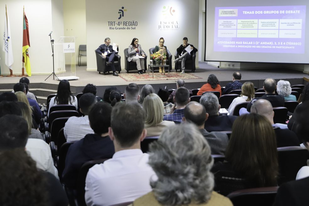 Público aparecem em primeiro plano, de costas, no auditório. Ao fundo, quatro pessoas estão sentadas em poltronas no palco, uma delas com o microfone na mão falando. Na esquerda aparecem bandeiras e na direita um telão ligado.