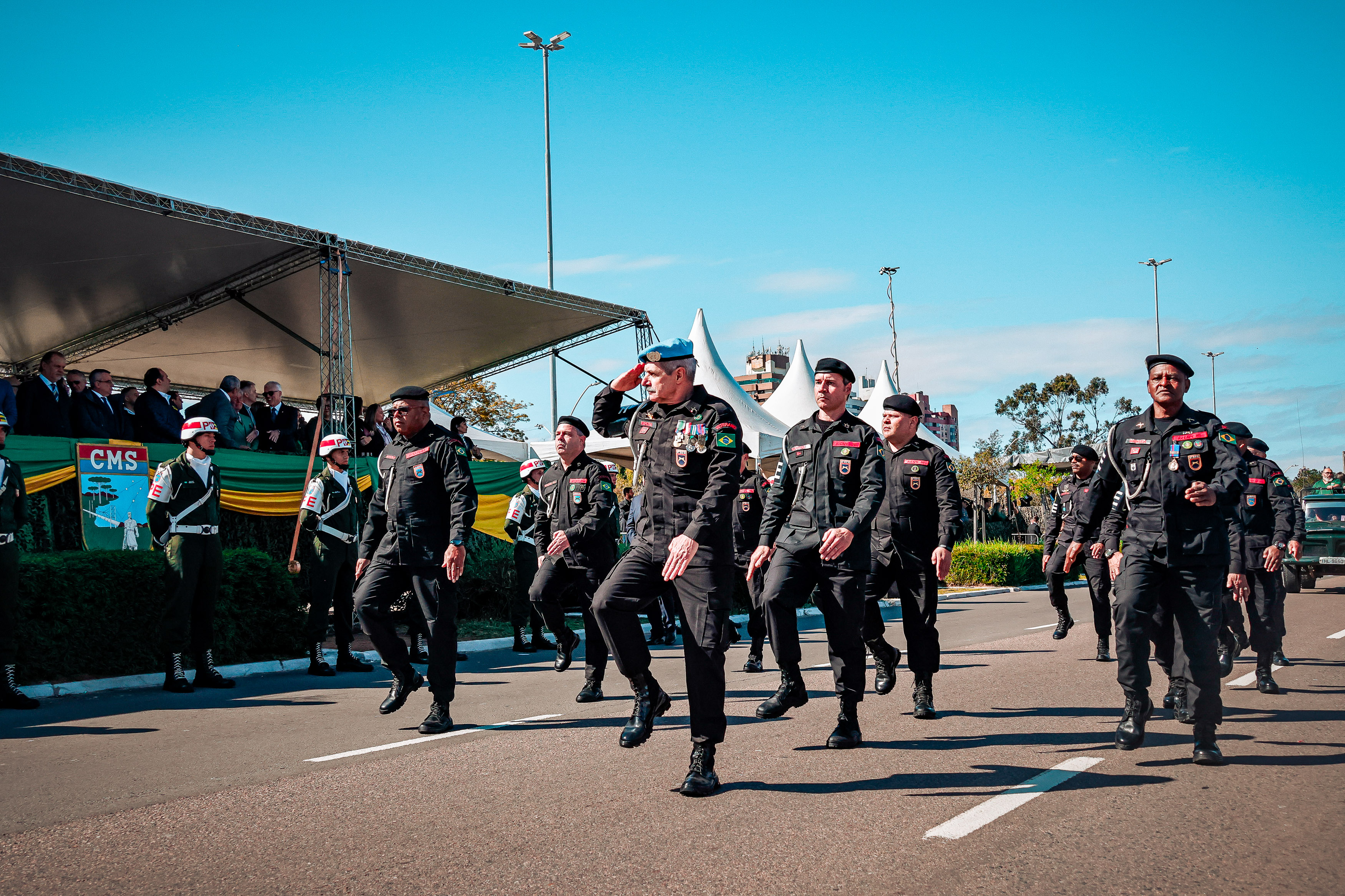Foto com plano aberto de pessoas marchando e um palanque ao fundo.