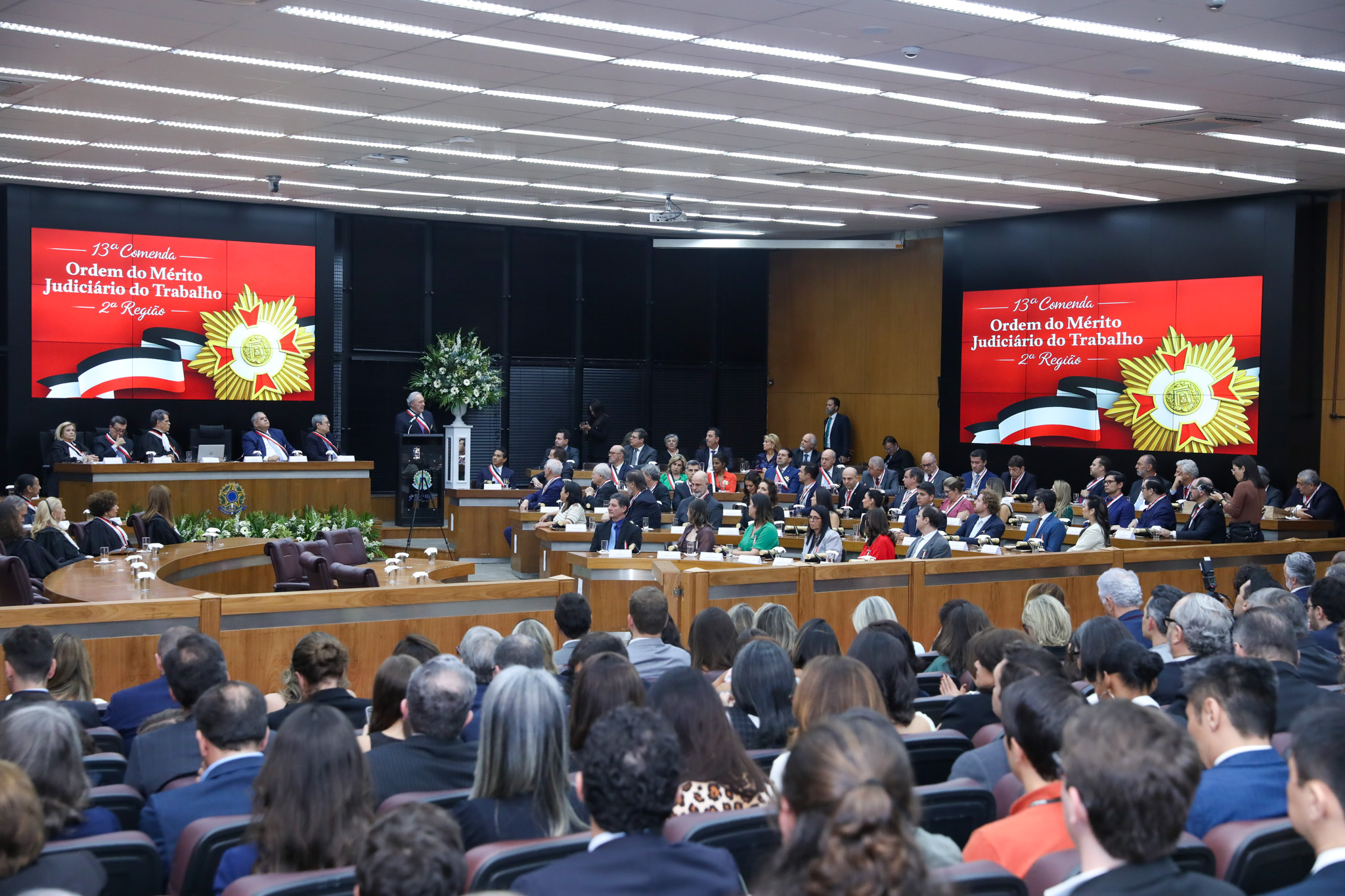 Foto em ângulo aberto da solenidade, mostrando público e mesa oficial.