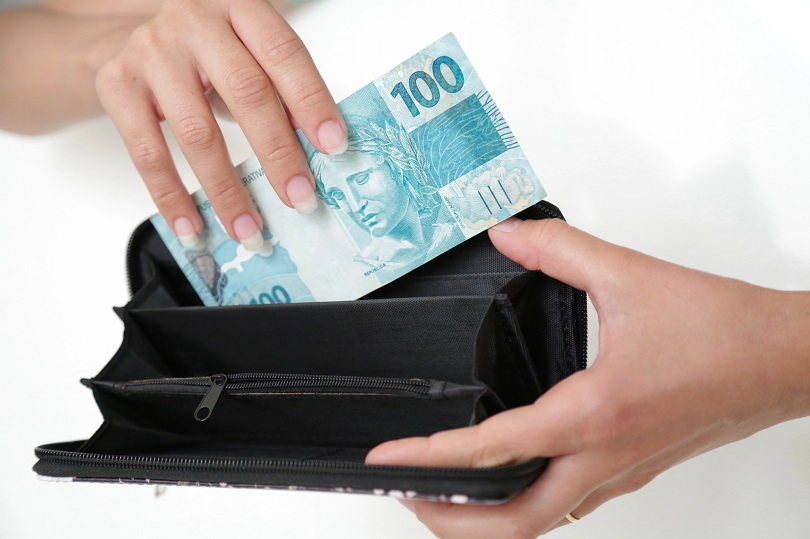 Foto ilustrativa mostra uma mão colocando uma cédula de 100 reais dentro de uma carteira preta aberta. A carteira está vazia.