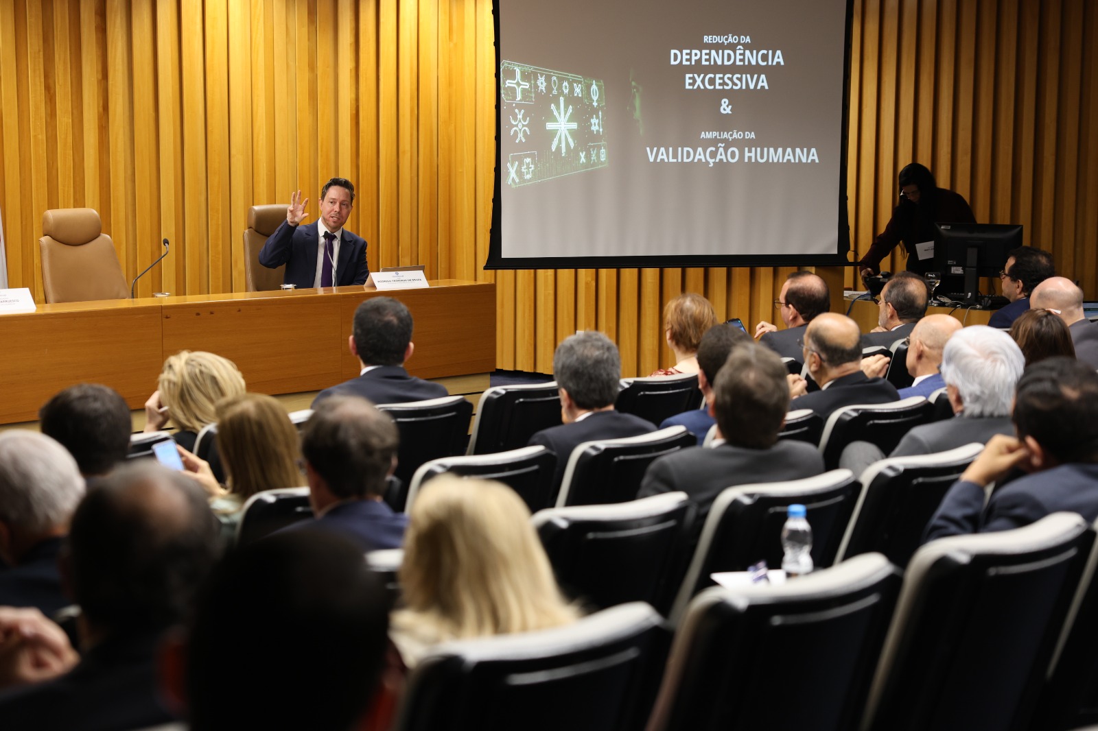 Palestrante de terno gesticula para a plateia em uma sala de conferências. Ao lado dele, o telão exibe o texto "REDUÇÃO DA DEPENDÊNCIA EXCESSIVA & AMPLIAÇÃO DA VALIDAÇÃO HUMANA".