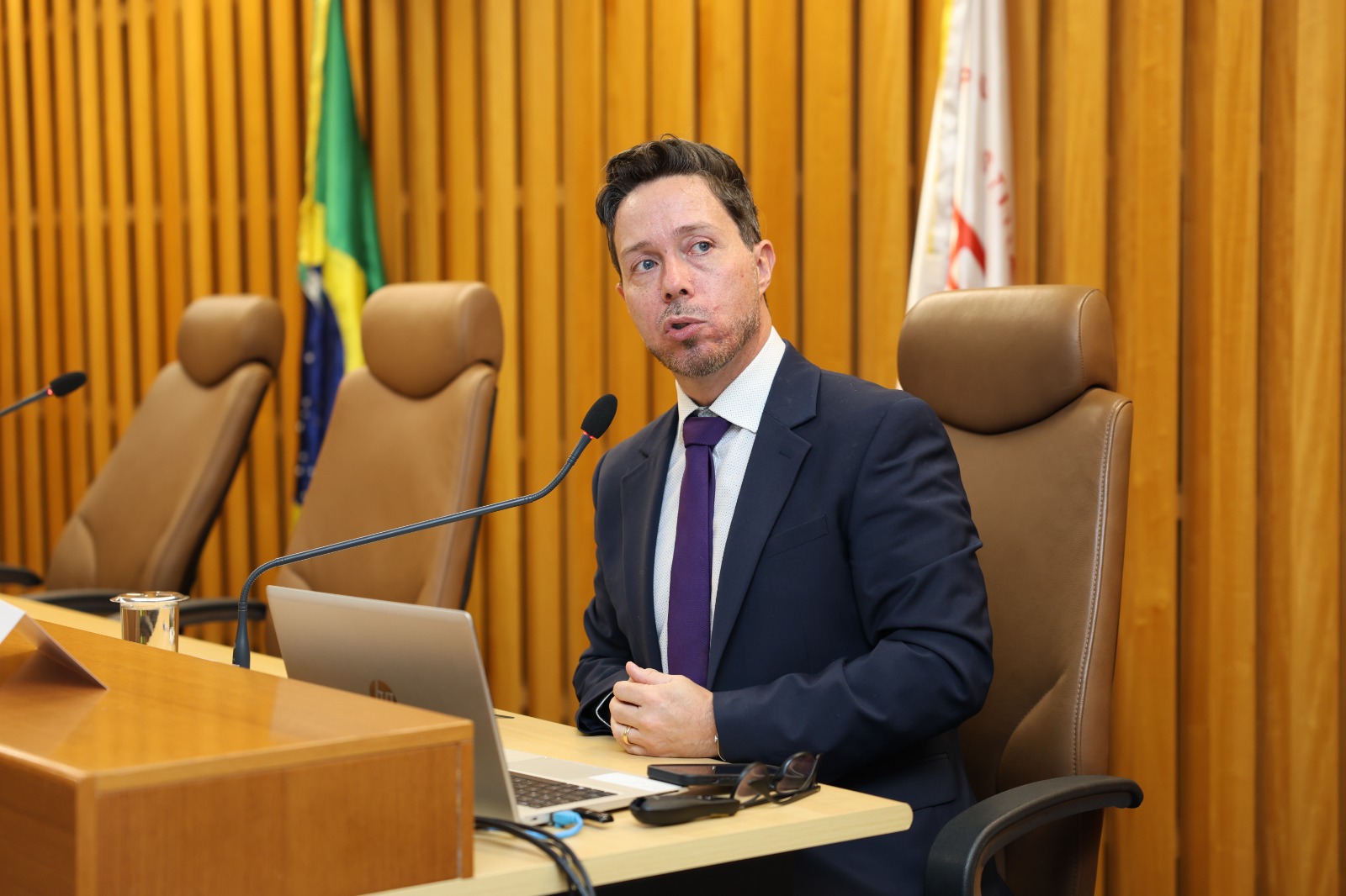 Rodrigo é branco e tem cabelo castanho curto. Usa terno azul e gravata roxa e discursa ao microfone em uma sala de audiências. Ao fundo, bandeiras, incluindo a do Brasil. Em frente ao plastrante, há um laptop sobre a mesa de madeira.