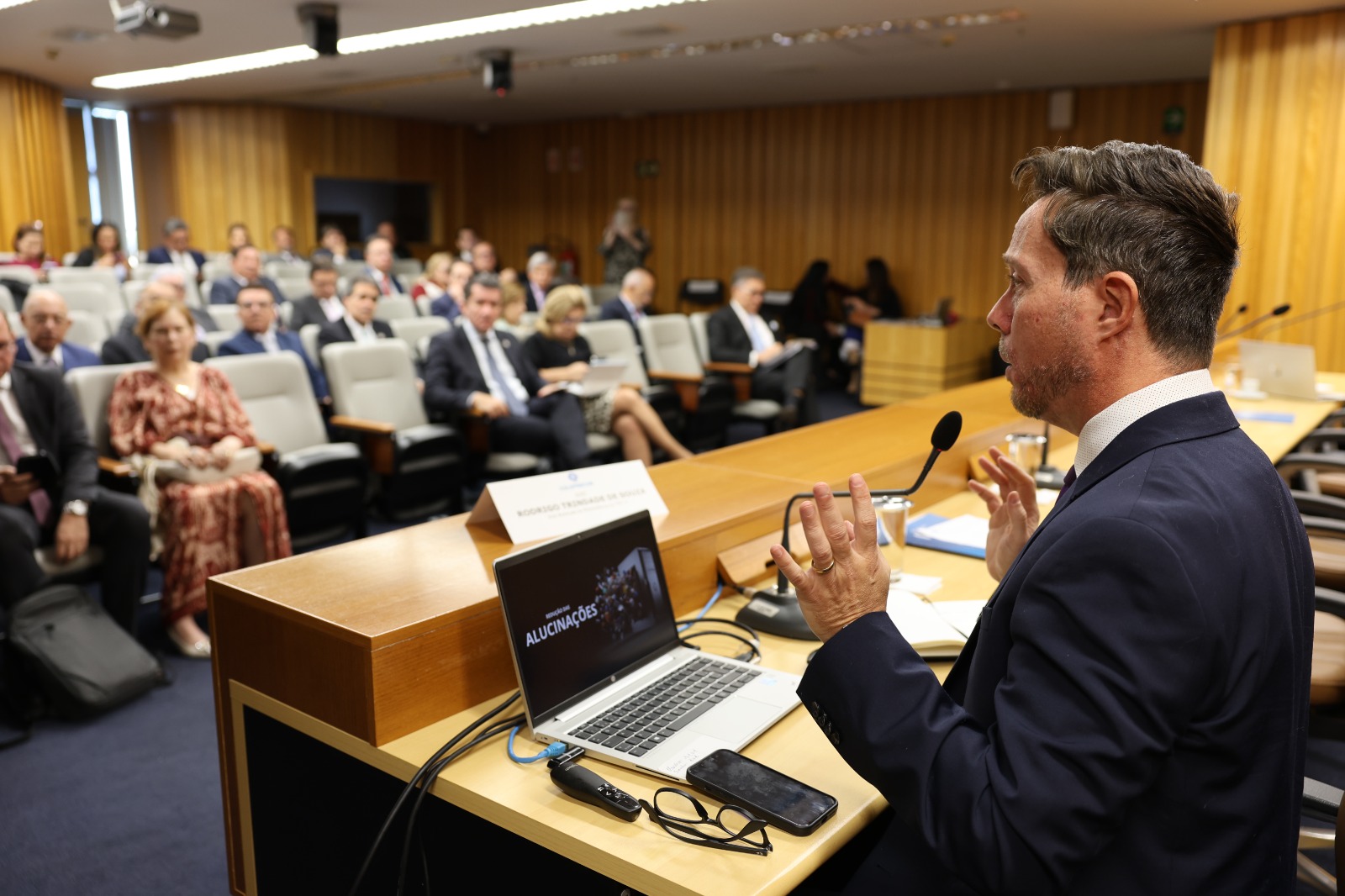 Homem de terno profere palestra em auditório. Ele está de perfil, falando e gesticulando, no canto direito da foto. Ao fundo aparece a plateia, fora de foco. Em frente ao palestrante, sobre a mesa, há um laptop aberto.