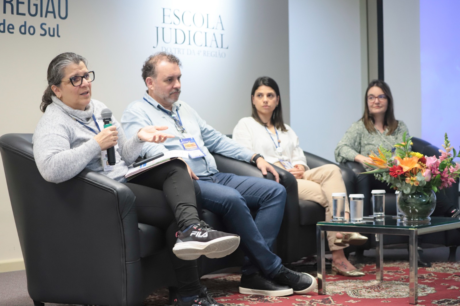 Quatro pessoas estão sentadas em poltronas no palco. Em primeiro plano, uma mulher fala ao microfone.
