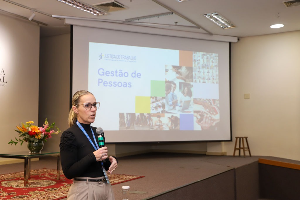 Maria Agusta fala de pé em primeiro plano. É branca, loira e usa óculos de aro grande. Ao fundo aparece um telão com o texto &quot;gestão de pessoas&quot;.