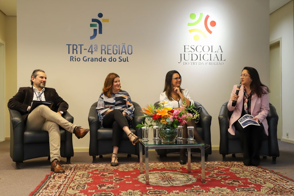 Quatro pessoas estão sentadas em poltronas no palco. A da direita está falando ao microfone. Todas sorriem. Na parede atrás aparecem os logos da JT e da Ejud4