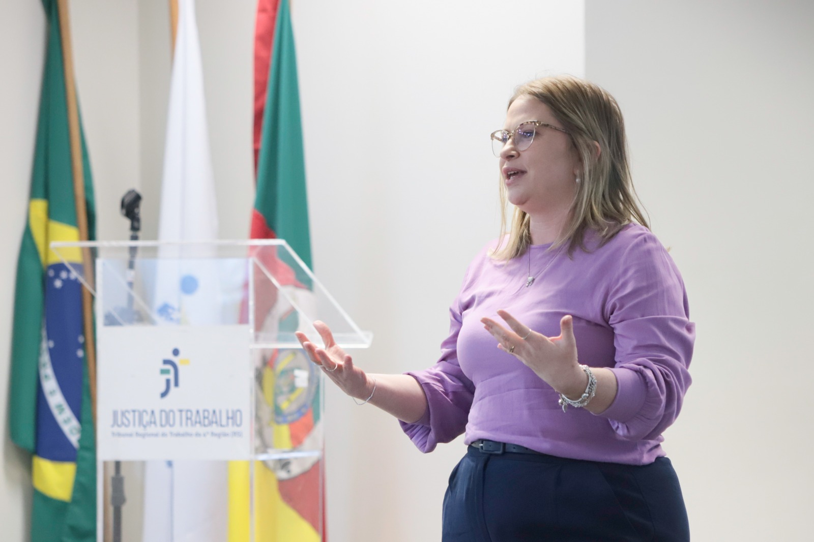 Caroline Vanzelotti é branca, loira e usa óculos de aro fino. Veste blusa roxa e calça preta. Está de pé, de perfil, falando e gesticulando. No lado esquerdo aparecem as bandeiras do Brasil, JT e RS, e uma bancada com o logo da JT.