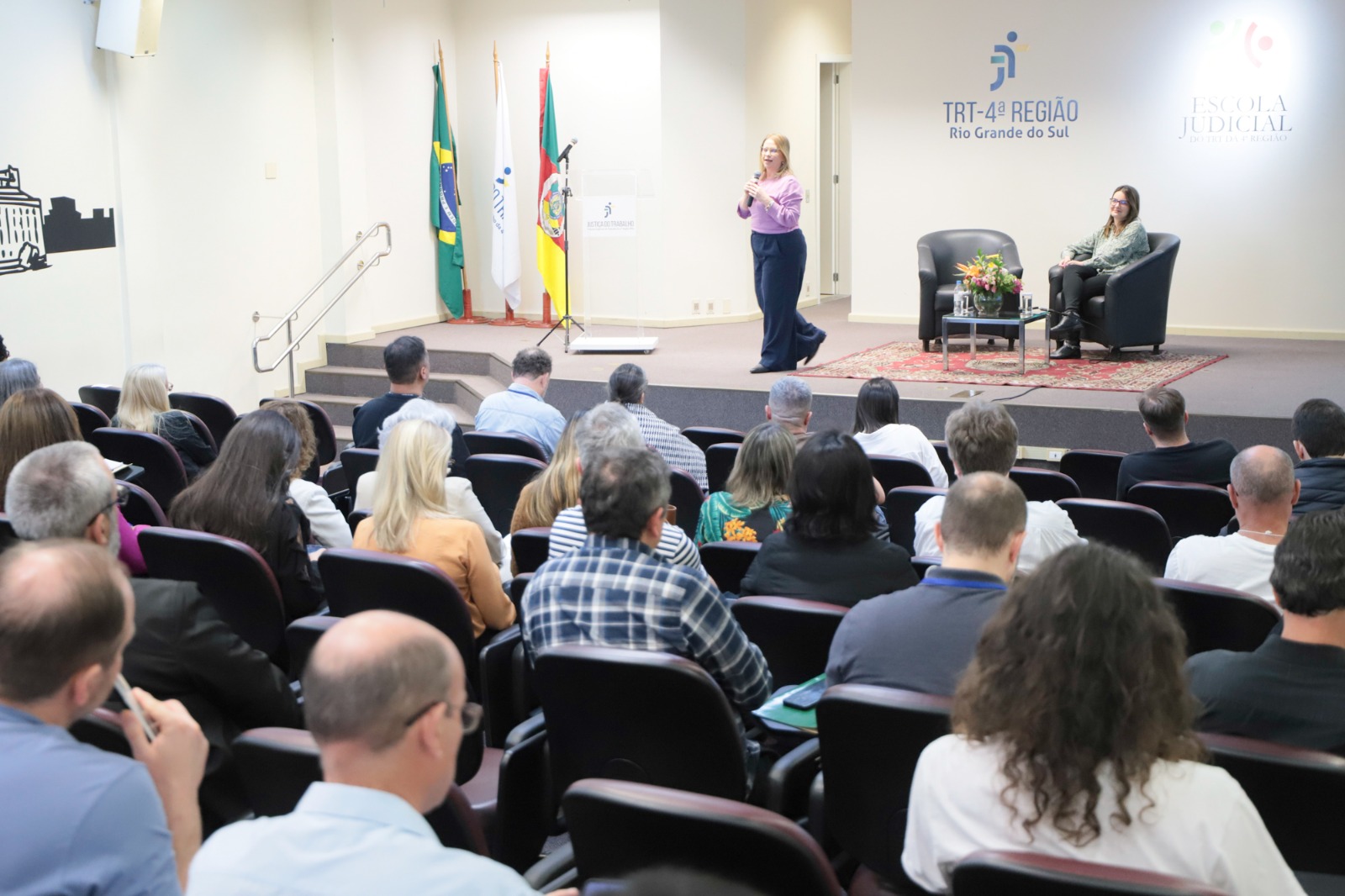 Público em primeiro plano. Ao fundo, no palco, aparecem duas mulheres. Uma está de pé, andando e falando ao microfone. A outra está sentada em um a poltrona assistindo.