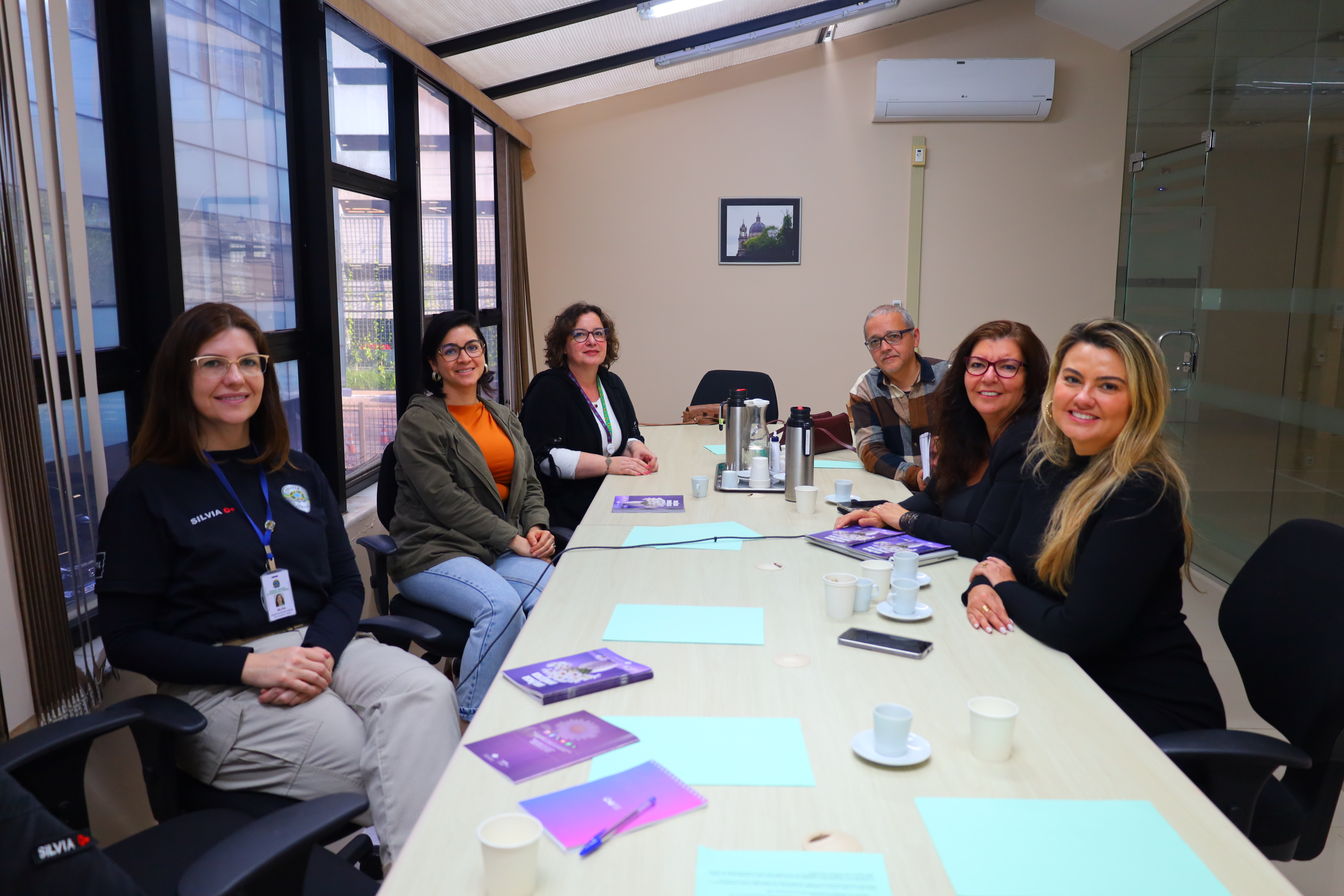 Seis pessoas sentadas em torno de uma mesa de reunião em sala clara, com janelas grandes à esquerda. Há cadernos e folhas sobre a mesa, além de xícaras e garrafas térmicas. Todos sorriem para a câmera.