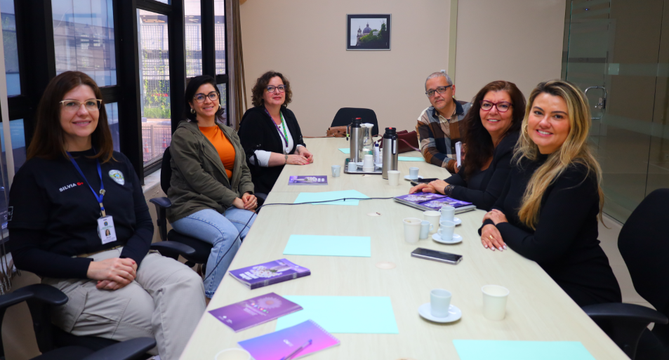 Seis pessoas sentadas em torno de uma mesa de reunião em sala clara, com janelas grandes à esquerda. Há cadernos e folhas sobre a mesa, além de xícaras e garrafas térmicas. Todos sorriem para a câmera.