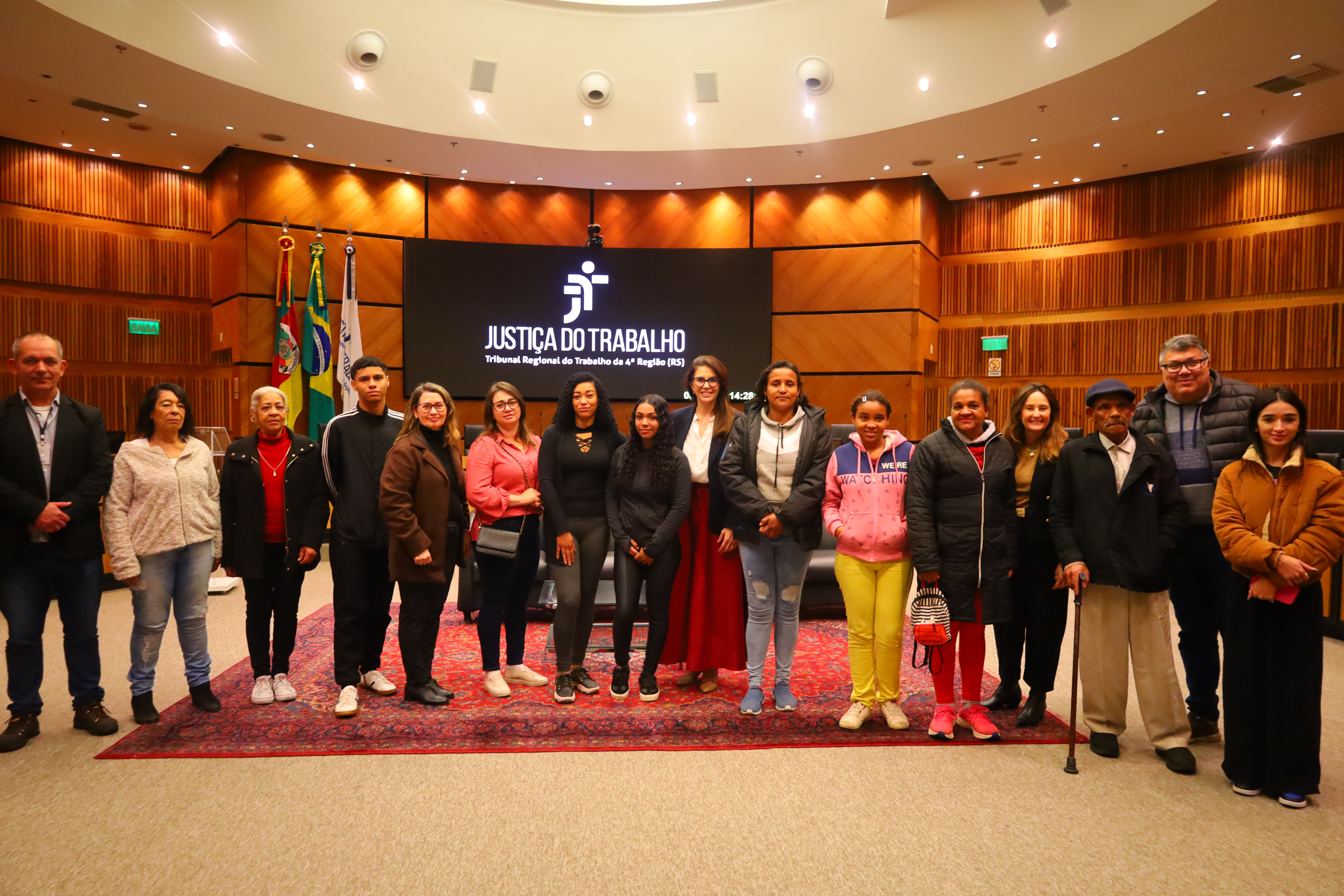 Fotografia dos estudantes no plenário em frente ao painel escrito “Justiça do Trabalho – Tribunal Regional do Trabalho da 4ª Região (RS)".
