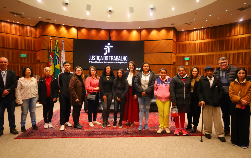 Fotografia dos estudantes no plenário em frente ao painel escrito “Justiça do Trabalho – Tribunal Regional do Trabalho da 4ª Região (RS)".
